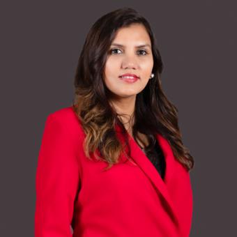 Dipali Trivedi in a red blazer, photographed against a dark grey studio backdrop.