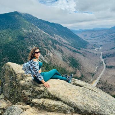 Dipali Trivedi resting on a granite ledge high above Crawford Notch, with Route 302 threading through the forested valley below