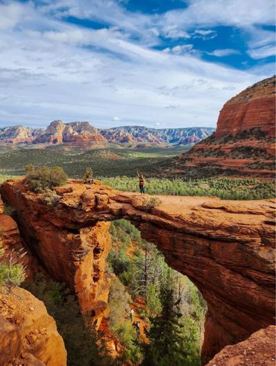 A hiker standing on the Devil's Bridge sandstone arch with an arm raised, Sedona's red rock formations stretching to the horizon
