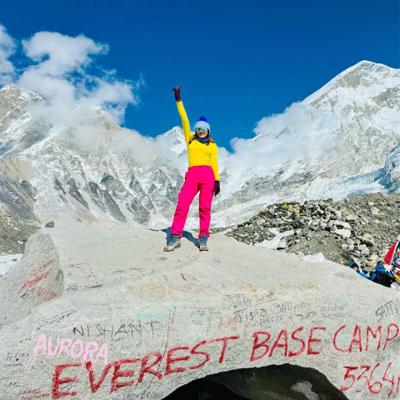 Dipali Trivedi standing with one arm raised on the Everest Base Camp boulder at 5,364 m, snow-covered Khumbu peaks rising behind her