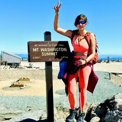 Dipali Trivedi beside the Mount Washington Summit sign marking 6,288 ft, trekking pole in hand under a clear blue sky