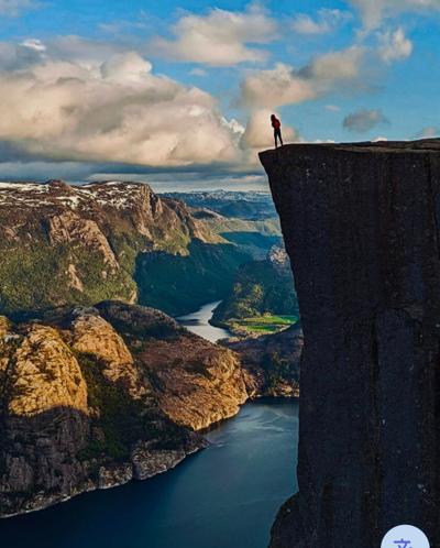 A lone hiker standing at the unrailed edge of the Preikestolen cliff plateau, the Lysefjord dropping away nearly 2,000 ft below