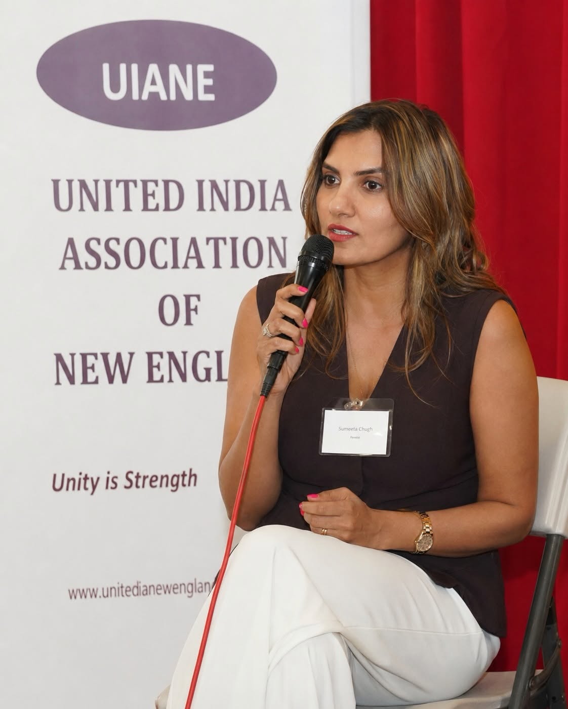 A panelist speaking into a microphone at the Youth Empowerment Summit, beside a United India Association of New England banner reading Unity is Strength